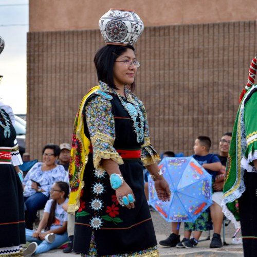 A woman in traditional attire walks gracefully, balancing a decorative pot on her head during a cultural event.