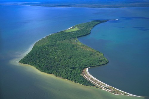 Aerial view of a lush green island extending into blue waters, surrounded by varying shades of sea.