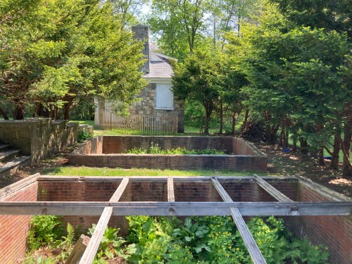 Ruins of a stone house surrounded by overgrown gardens and trees, with visible brick foundations and wooden beams.