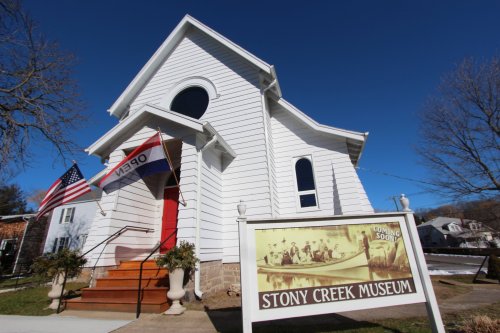 Exterior of Stony Creek Museum, featuring a white building, red door, flags, and a sign in front. Clear blue sky above.