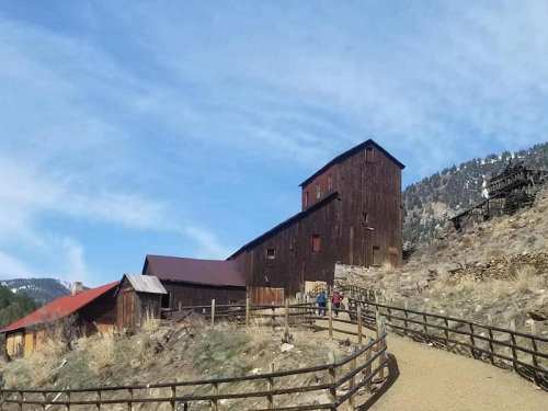 A rustic wooden barn on a hillside, with a dirt path and a wooden fence, under a blue sky with scattered clouds.