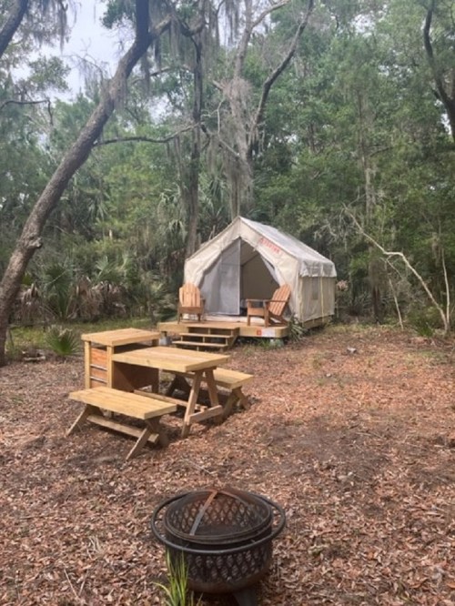 A glamping tent surrounded by trees, with picnic tables and a fire pit in a natural setting.