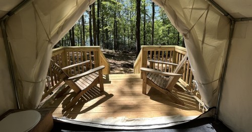 View from inside a tent, showcasing a wooden deck with two chairs and a forest backdrop.