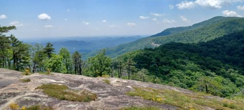 A panoramic view of lush green mountains under a clear blue sky, with rocky terrain in the foreground.