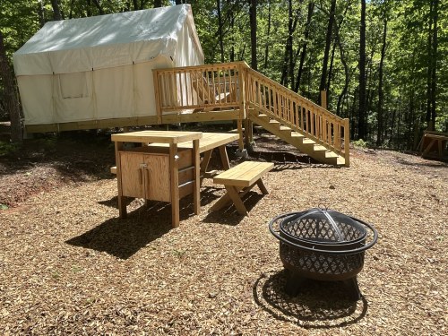 A wooden deck with a picnic table and fire pit in front of a tent cabin, surrounded by trees.