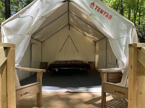 A spacious white tent interior with a bed and wooden chairs on a deck, surrounded by trees.