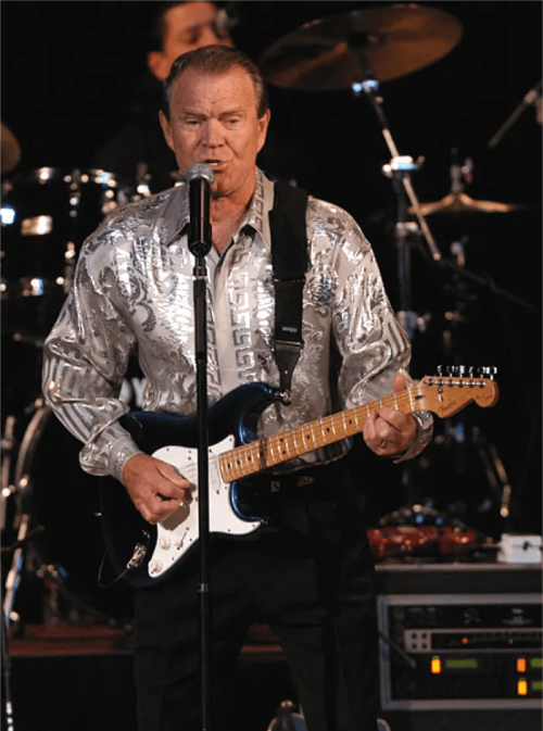 A musician performs on stage with a guitar, wearing a shiny silver shirt, with a drum set in the background.