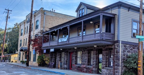 Historic buildings with a wooden balcony and stone facade, set on a quiet street with trees and power lines.