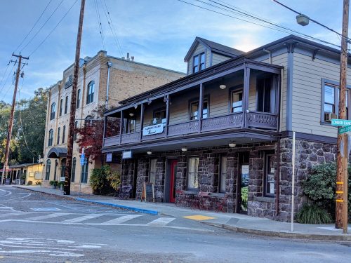 Historic buildings with a balcony and stone facade, set on a street corner with power lines and trees nearby.
