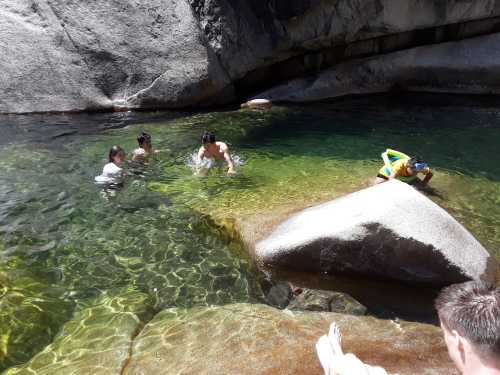 Four people swimming in a clear, greenish pool surrounded by large rocks and sunlight. One person is on a float.