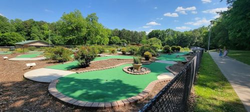 A sunny mini-golf course with green turf, surrounded by trees and a walking path in the background.