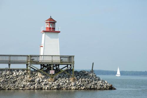 A white lighthouse with a red roof stands on a rocky pier, with a sailboat in the background on calm waters.