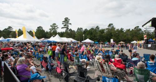 A large crowd gathers at an outdoor festival, seated in chairs with tents and trees in the background under a cloudy sky.