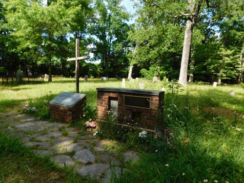 A peaceful cemetery scene with a cross, a brick memorial, and overgrown grass surrounded by trees and gravestones.