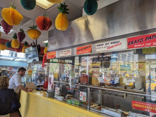 A colorful fast-food counter with pineapple decor, featuring a menu and customers ordering at Gray's Papaya.
