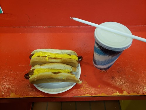 Two hot dogs with melted cheese on a paper plate, next to a cup with a straw on a red table.
