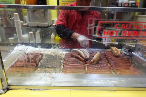 A vendor prepares hot dogs in a display case, with several cooked hot dogs and buns visible.