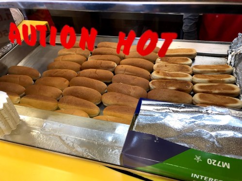 A display of freshly steamed hot dog buns in a food cart, with a "Caution Hot" sign in red.