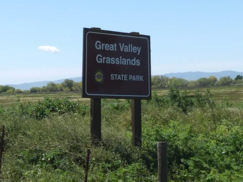 Sign for Great Valley Grasslands State Park, surrounded by greenery and distant mountains under a clear blue sky.