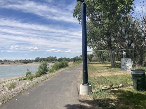 A pathway along a river with a sign warning about stray golf balls near a golf course area. Trees and blue sky in the background.