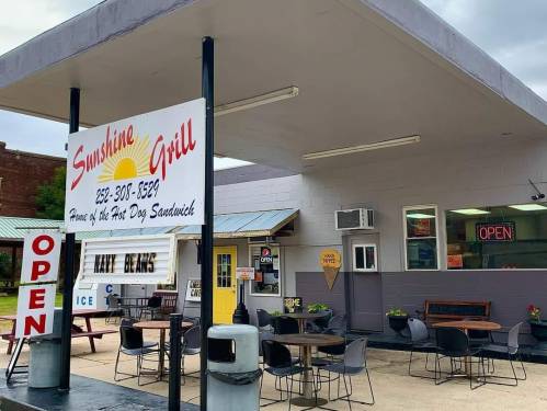 Exterior of Sunshine Grill with a sign, outdoor seating, and an "Open" neon sign. Cloudy sky in the background.