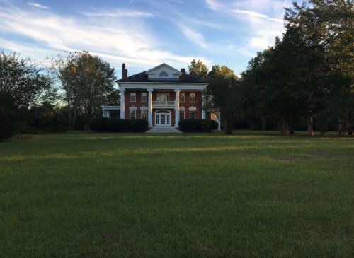 A large, two-story brick house with white columns, surrounded by green grass and trees under a blue sky.