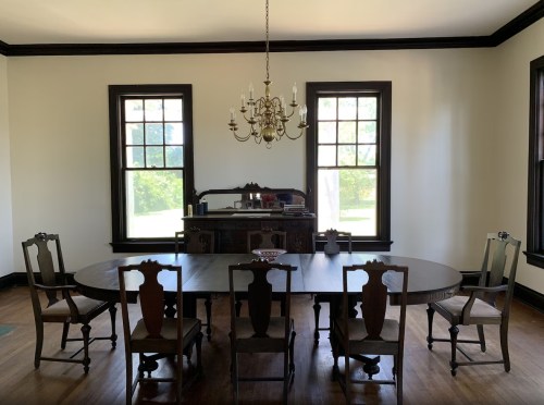 A spacious dining room featuring a large oval table, six chairs, a chandelier, and two windows with natural light.