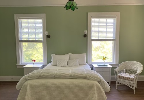 A cozy bedroom with a green wall, a white bed, two windows, and a wicker chair beside a nightstand with flowers.