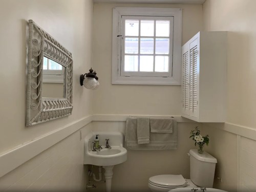 A small, bright bathroom featuring a white mirror, sink, toilet, and a window with natural light.