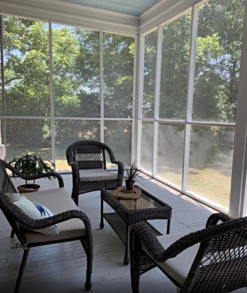 A cozy screened porch with wicker chairs, a small table, and greenery visible through the screens.