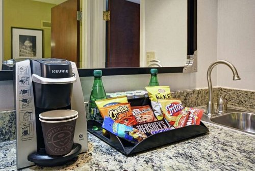 A hotel room coffee station with a Keurig, snacks, and drinks on a granite countertop.