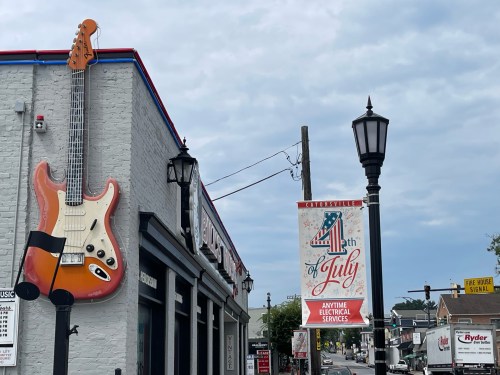 A large guitar mural on a building with a "4th of July" banner and a street lamp in a small town setting.
