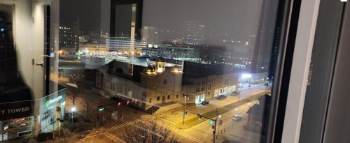 Nighttime cityscape view from a window, featuring illuminated streets and buildings under a cloudy sky.
