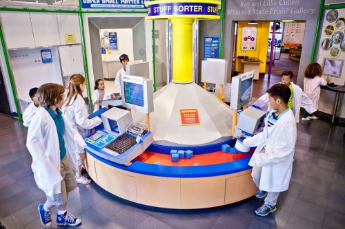 Children in lab coats interact with a colorful sorting exhibit in a science museum, exploring materials and learning.