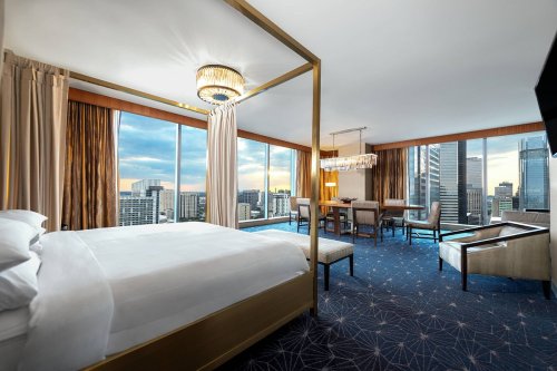 Luxurious hotel room with a four-poster bed, large windows, and modern furnishings overlooking a city skyline.
