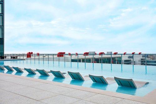 Rooftop infinity pool with lounge chairs, overlooking a city skyline under a cloudy sky.