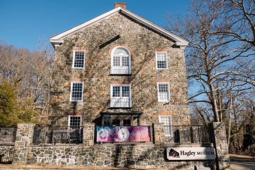 Stone building of Hagley Museum with a sign, surrounded by trees and a clear blue sky.
