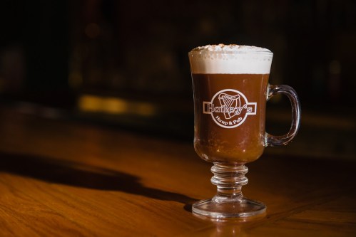 A glass of coffee topped with whipped cream and a sprinkle of cinnamon, sitting on a wooden table.
