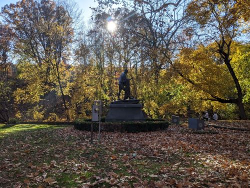 A statue in a park surrounded by colorful autumn trees and fallen leaves, with sunlight filtering through the branches.