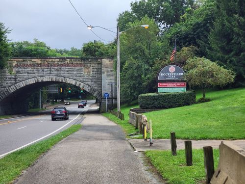 A road leads under a stone bridge, with a sign for Rockefeller Park and greenery on either side.