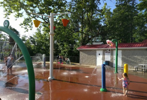 Children play in a splash pad with colorful water features on a sunny day, surrounded by trees and a gray building.