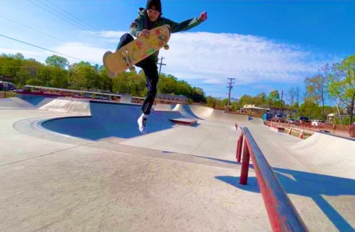 A skateboarder performs a jump over a rail at a skatepark on a sunny day.