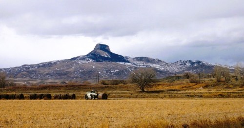 A scenic landscape featuring views of Heart Mountain in Wyoming dusted with snow, and a field with hay bales under a cloudy sky.