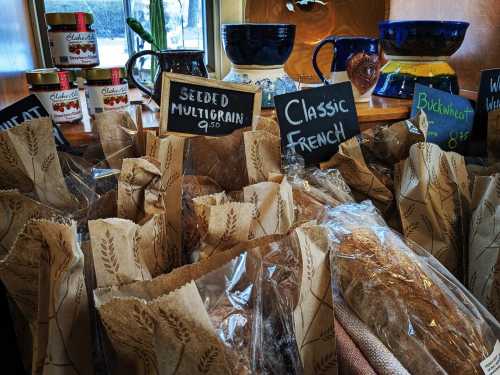 A display of various bread loaves in paper bags, labeled with prices, in a cozy shop setting.