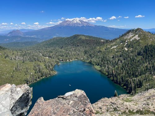 A scenic view of a blue lake surrounded by lush greenery, with a snow-capped mountain in the background under a clear sky.