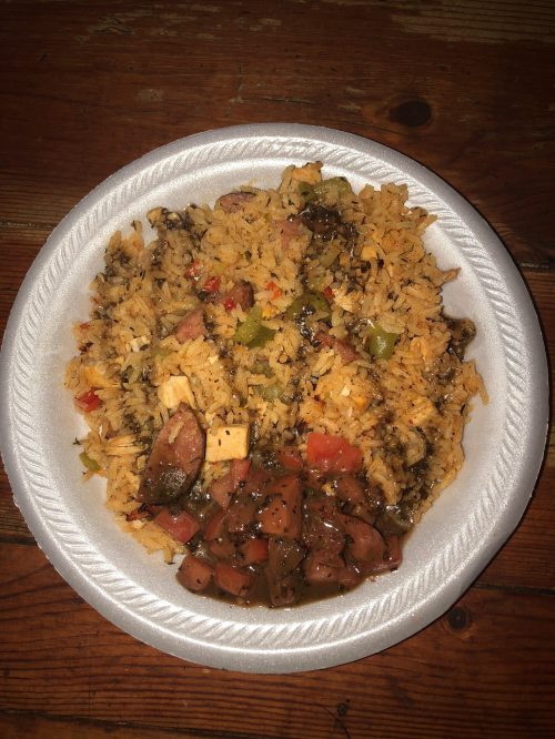 A plate of seasoned rice mixed with vegetables and chunks of sausage, served on a disposable white plate.