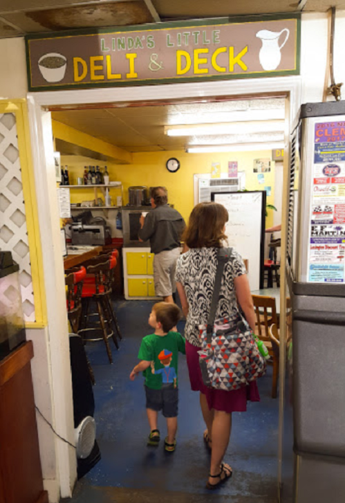 A woman and child enter "Linda's Little Deli & Deck," a cozy eatery with colorful decor and a welcoming atmosphere.