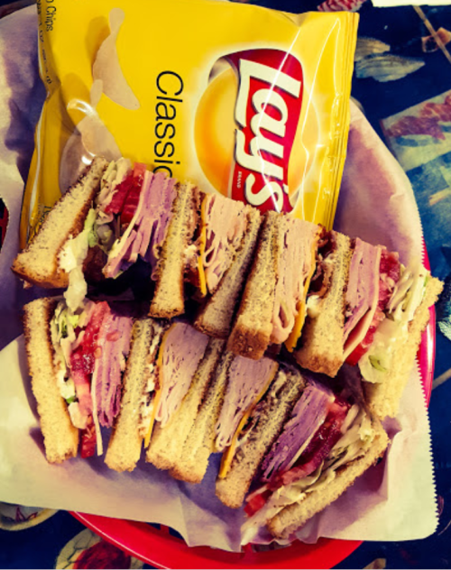 A plate with a stacked sandwich filled with meats and veggies, alongside a bag of Lay's Classic potato chips.