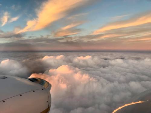 Aerial view of fluffy clouds illuminated by a colorful sunset, with a plane's wing and engine in the foreground.