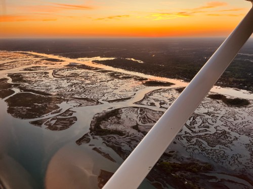 Aerial view of winding waterways and marshlands at sunset, with vibrant orange and yellow hues in the sky.
